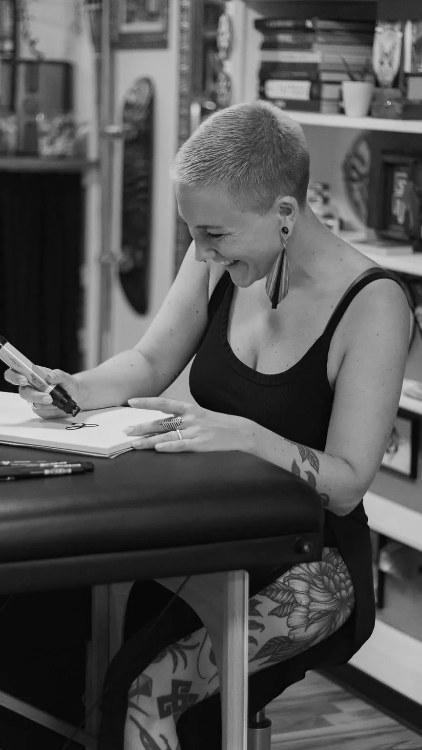 Kaiiza assise à la table du studio dessine dans un carnet, souriante, bibliothèque en arrière-plan
