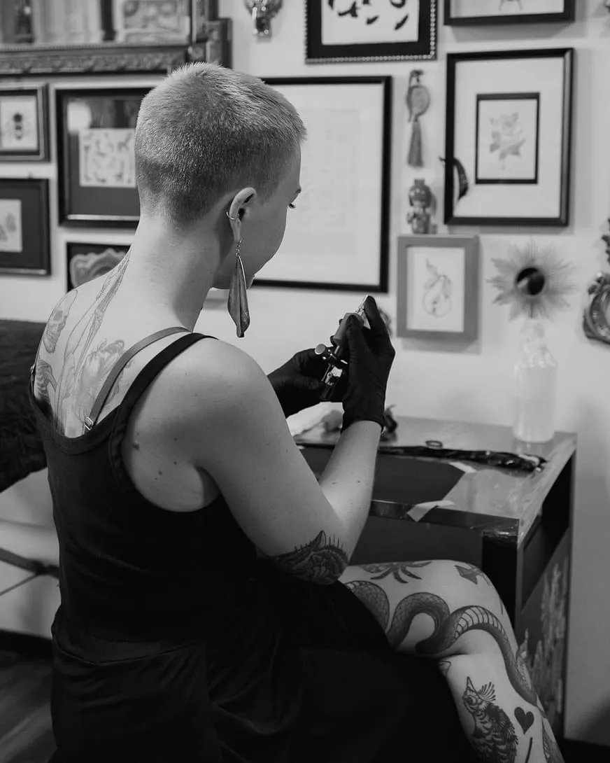Kaiiza vue de dos, assise à son poste de travail, prépare la machine à tatouer devant le mur de cadres