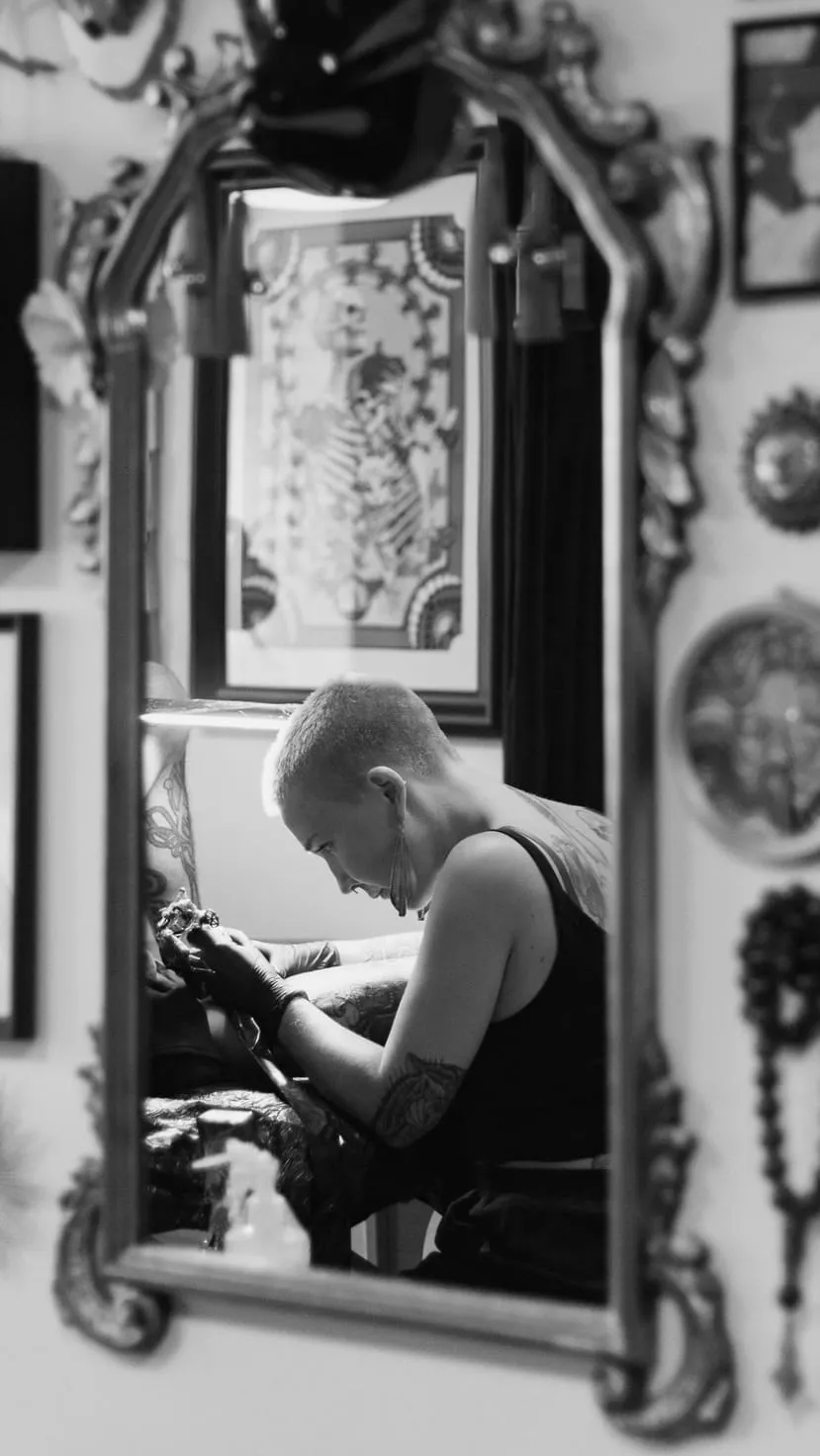 Kaiiza en séance de tatouage, vue dans le reflet d'un grand miroir au cadre baroque du studio.