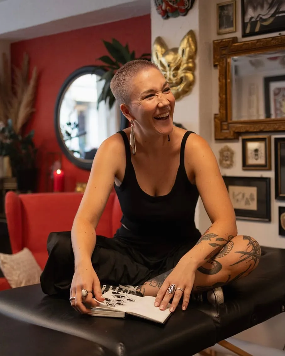 Kaiiza assise en tailleur sur la table de tatouage rit, carnet ouvert, devant le mur rouge et les cadres dorés du studio.