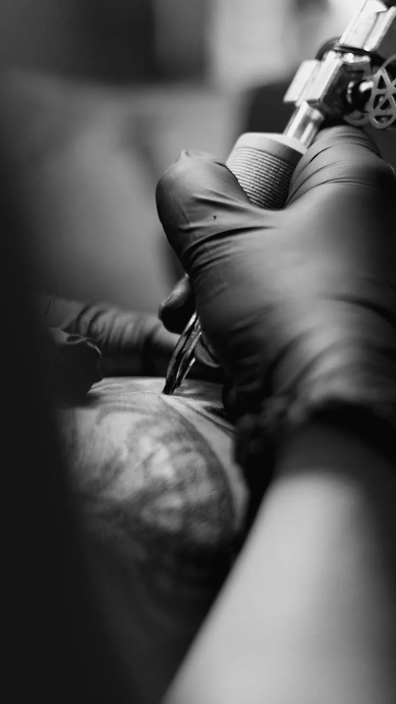 Plan serré vertical sur la machine à tatouer tenue par une main gantée de noir au contact de la peau