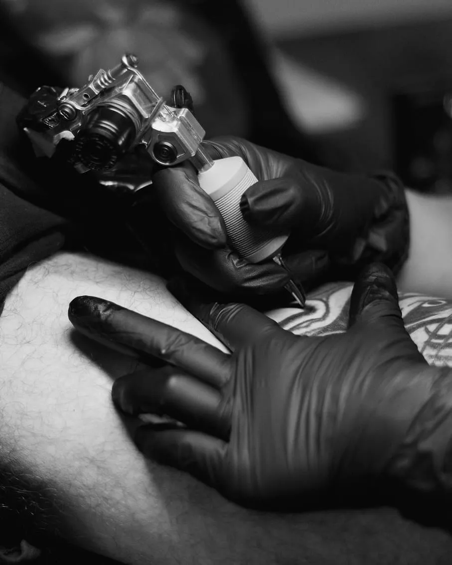 Gros plan sur la machine à tatouer posée sur la peau, mains gantées de noir tendant la zone.