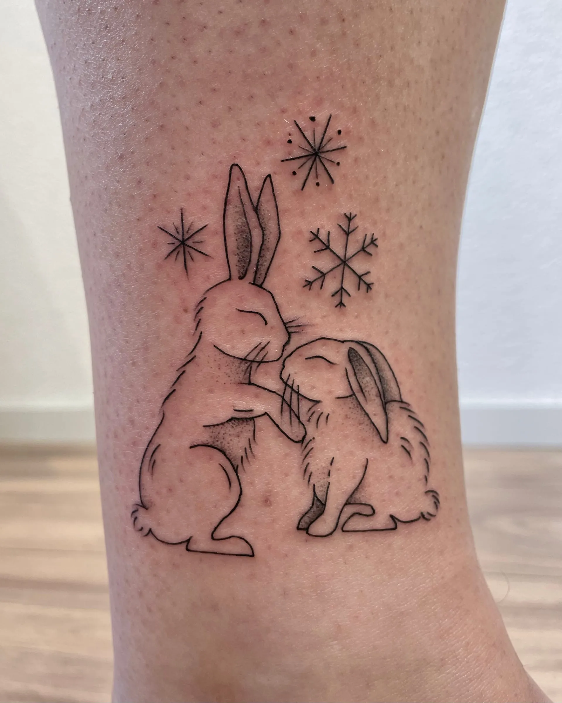 Tatouage fineline de deux lapins qui se cajolent entourés de flocons de neige, sur la cheville.