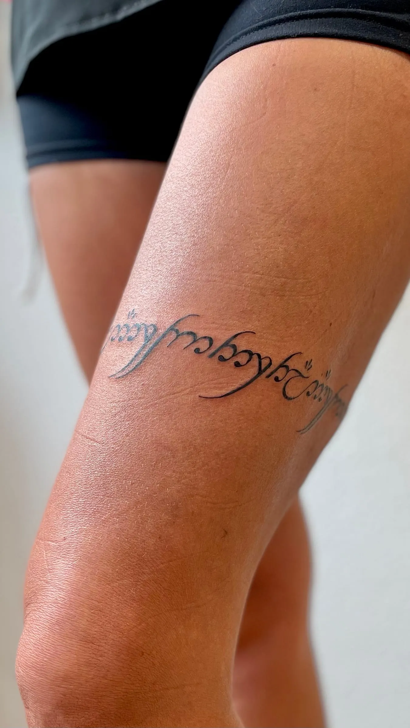 Tattoo in fine line of a phrase in cursive Elvish script on the side of the thigh.