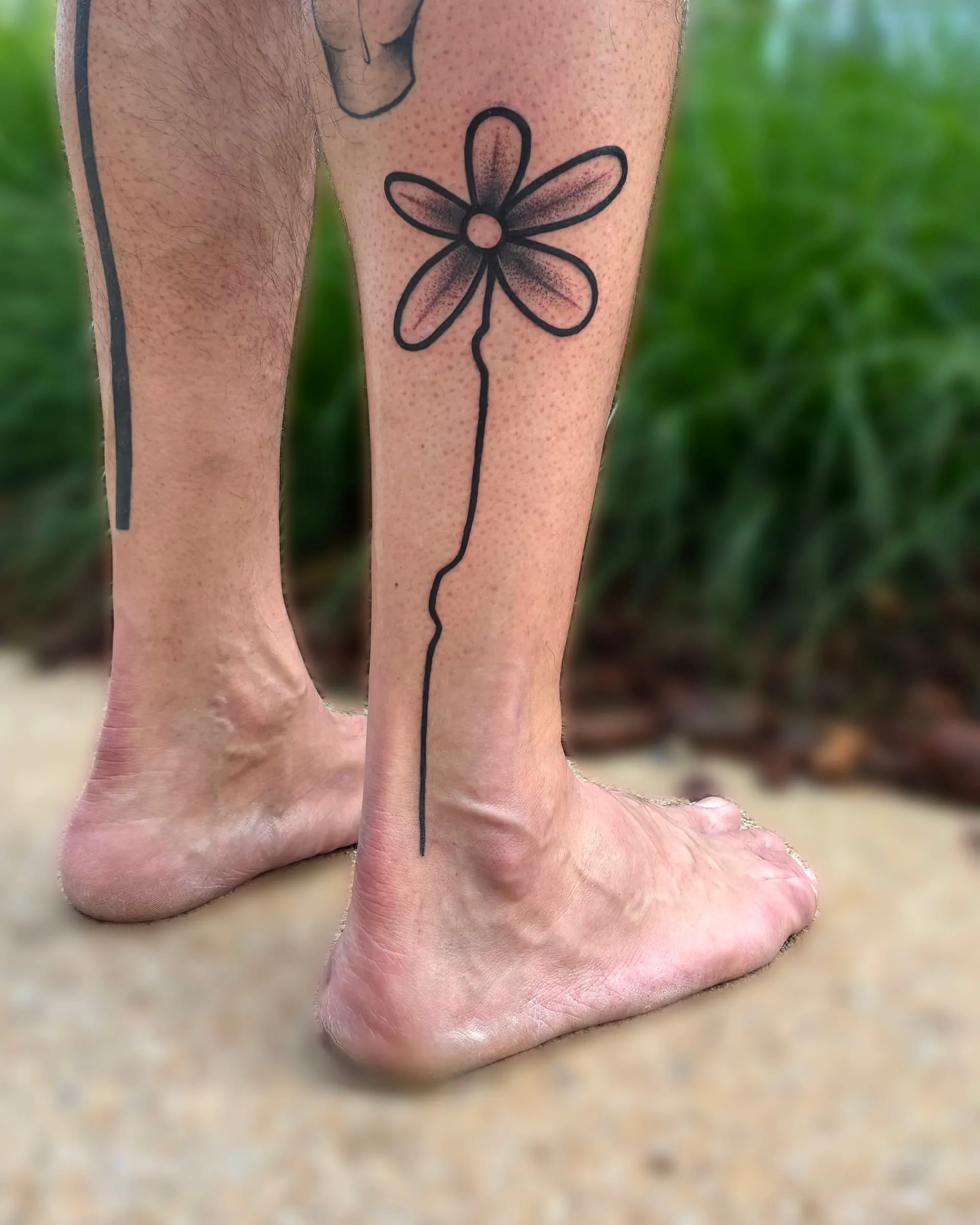 Tatouage en ligne d'une fleur stylisée dont la longue tige descend le long du mollet jusqu'à la cheville