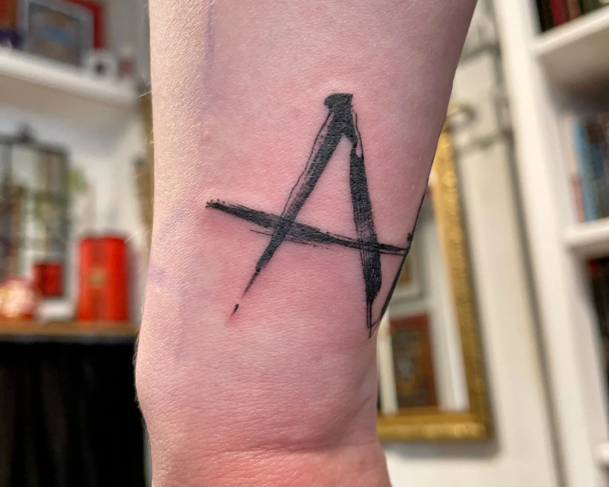 Tatouage d'une lettre A tracée en larges coups de pinceau noirs, sur l'avant-bras.