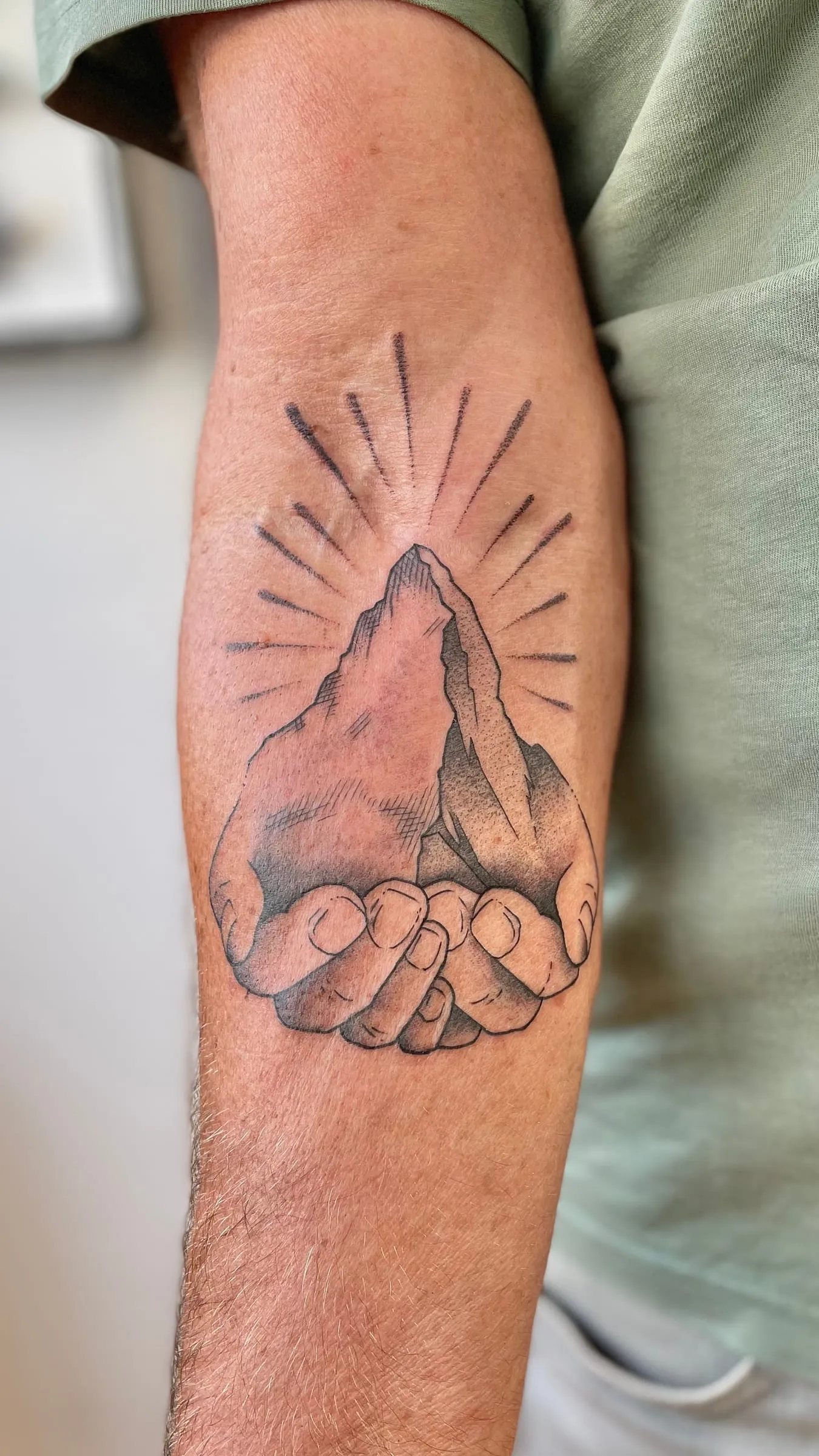 Tatouage en noir et gris de deux mains jointes tenant une montagne surmontee de rayons, sur l'avant-bras.