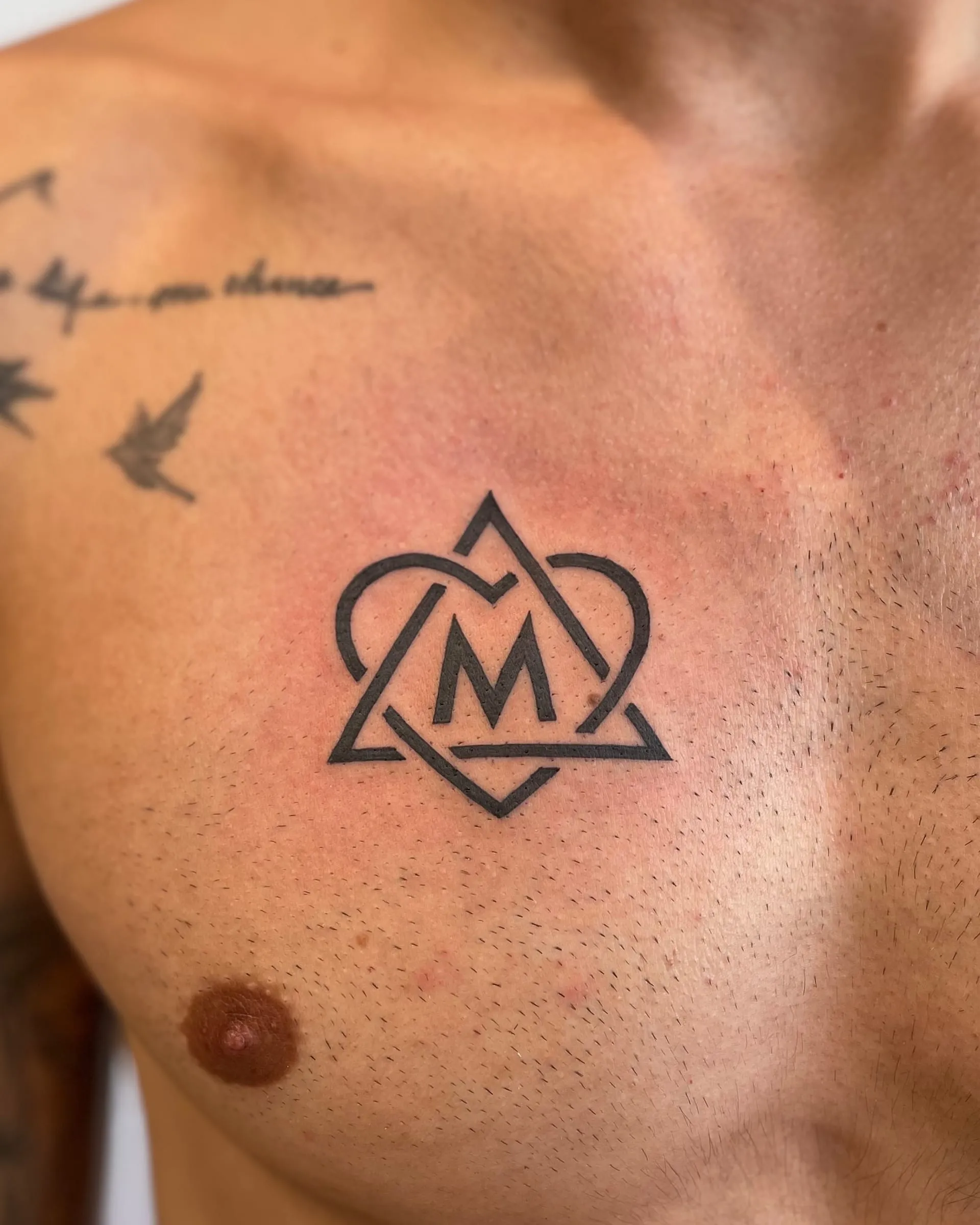Tatouage graphique d'un monogramme M encadre par un coeur et un triangle, sur le torse