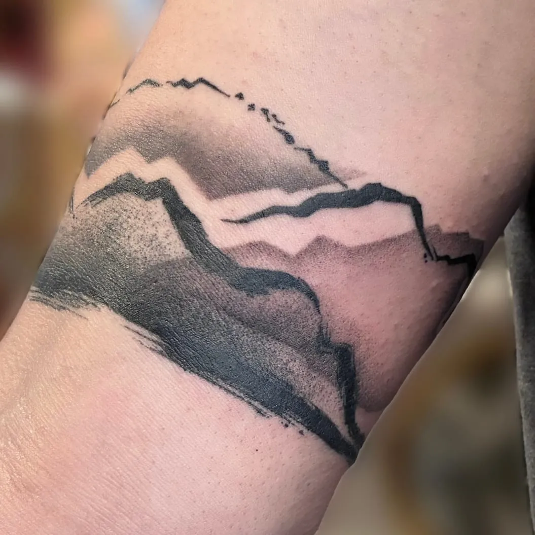 Tatouage en noir et gris d'une chaîne de montagnes en encre brossée, formant un bracelet autour du bras