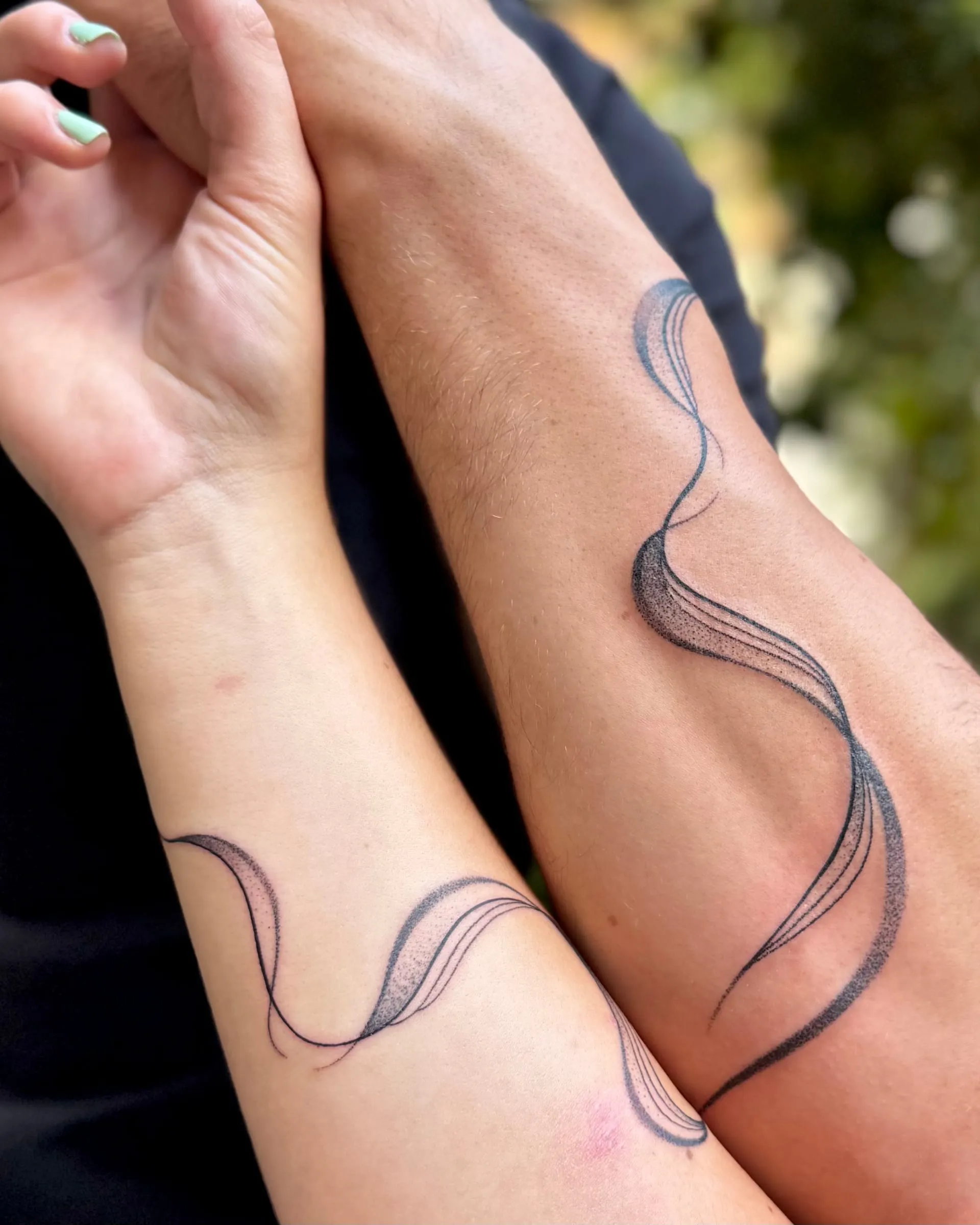 Tatouages assortis de rubans noirs ondulés sur deux avant-bras rapprochés, contours fins et ombrages en pointillés